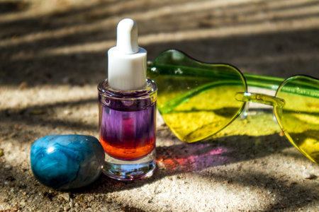 Glass cosmetic bottle with purple-red liquid oil on sandy shore in summer. Care cosmetics for women. Serum jar on natural background. green sunglassesの写真素材