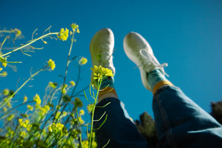 Woman's feet in white sneakers raised up against the blue sky. A girl resting on a spring meadow among blooming Yellow mustard flowers in spring day.の写真素材