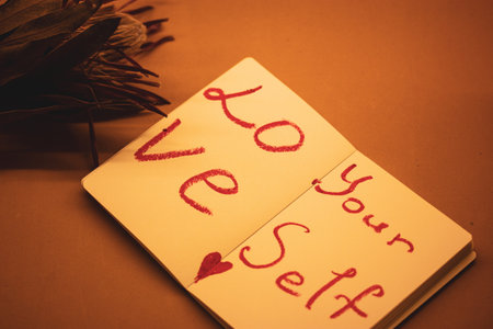 Text 'Love yourself' written in red lipstick on the sheets of open paper notebook on orange background. Self-love, healthy narcissism, motivation.の写真素材