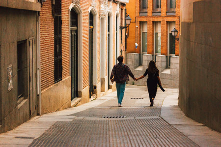 Madrid, Spain. 1 May 2024 A couple of woman and man are walking down an old city street in sunny day holding by the hands. Romantic people traveling.のeditorial素材