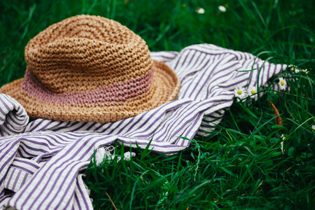 Female boho style straw hat lies on a striped plaid on a green grass lawn of blooming meadow. Summertime journey, travel. Summer vacation in nature.の写真素材