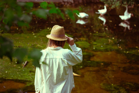 Young woman in a straw sun hat stands on pond, lake shore overgrown with greenery in a summer garden, a park. Rural farm people lifestyle. White Geeseの写真素材