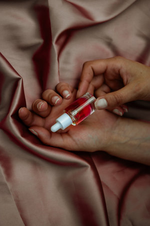 Transparent glass pipette bottle with red serum for face, body care in the hands on silky pink textile. Female beauty treatment. Beautician productの写真素材