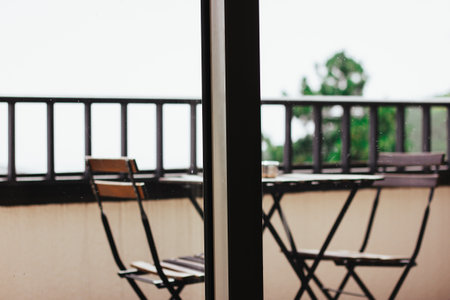 Wooden table and chairs on balcony behind a panoramic window. Garden furniture at home. Empty patio in a house in a forest. Railing on terrace.の写真素材