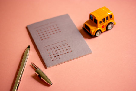 Toy yellow American school bus, calendar with dates, golden pen on pink table. Stationery for...