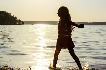 Teenage girl silhouette actively walking along the bank of river, lake at sunset in summer. Recreation season in children's camp. Sailboat in sunlightの写真素材