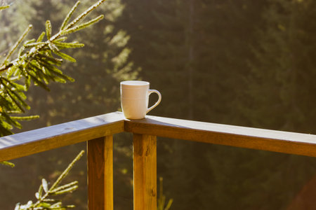 White ceramic cup of tea, coffee beverage stands on wooden balcony railing against green coniferous forest background, tall pines, firs in woods. Breakfast in the mountains. Spruce branch in sunlight.の写真素材