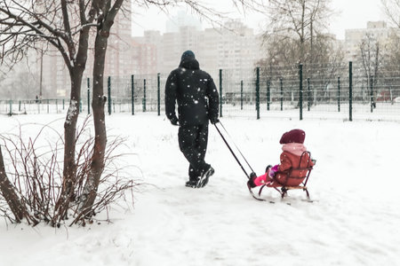 A father pulls sled with a child on a frosty cold winter day. A walk with a little kid in winter season. Falling snowflakes in urban scene. Family funの写真素材