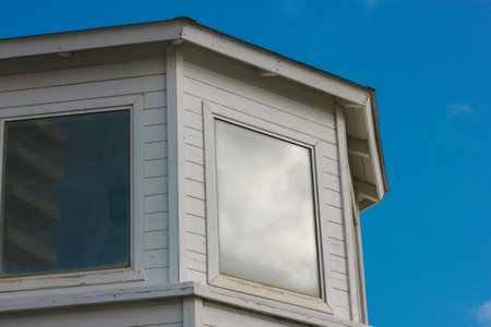 Bay windows. Facade of a rural white wooden house with two windows in the daytime. Corner of an old abandoned building against blue sky. Flat roofの写真素材