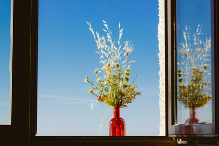 Authentic still life with red vase with wildflowers on windowsill on background of open window in...