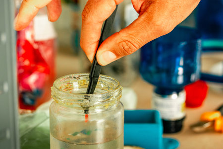 A person's hand stirs a substance with a tool in a glass jar, showcasing a close-up view with tools...
