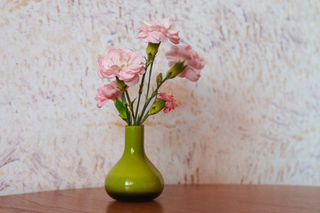 A beautiful still life featuring pink carnations in a green vase set on a wooden table, with a patterned wall behind. A delightful tabletop setting.の写真素材