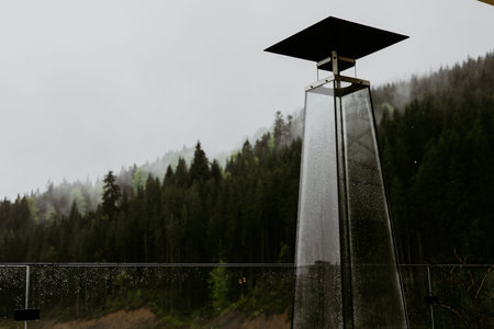A sleek, modern glass heater on a misty mountain day, with lush green trees in background on a rainy...
