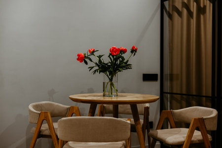 A beautiful arrangement of red peonies sits gracefully on a wooden table with accompanying chairs. A...