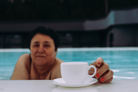 A relaxed woman enjoying a cup of coffee while relaxing in a refreshing swimming pool. A woman in a...