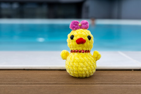 A cute, handmade crochet chick wearing a pink bow sits by a pool. Summertime vacation. A funky...
