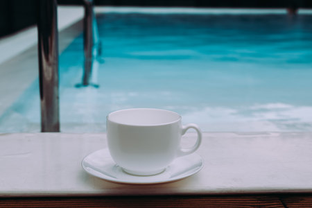 A white teacup sits beside a tranquil blue pool, a peaceful scene. A serene scene featuring a white teacup and saucer set by a pool, evoking relaxation and tranquility.の写真素材