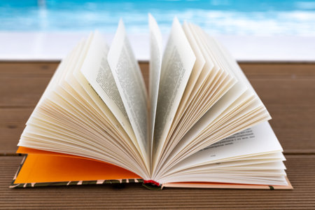 An inviting open paper book sits on a wooden deck near a pool, ready for summer reading enjoyment. Turning the pages of a book on summer vacation.の写真素材
