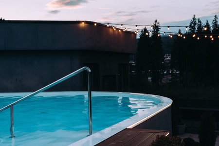A rooftop pool scene at twilight, adorned with string lights, creating a relaxing and tranquil atmosphere. A serene evening view of a rooftop pool, string lights, and a backdrop of trees.の写真素材