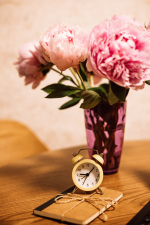 Beautiful pink peonies in a purple vase with a vintage alarm clock and a notebook. A delicate bouquet of pink spring flowers on a wooden table at homeの写真素材