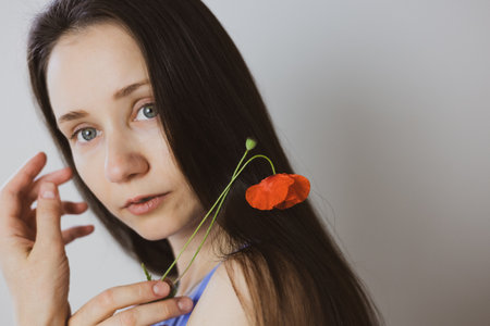 A beautiful brunette young white woman gazes at the viewer, with a red poppy gently resting in her dark hair. Natural female beauty in summertime.の写真素材