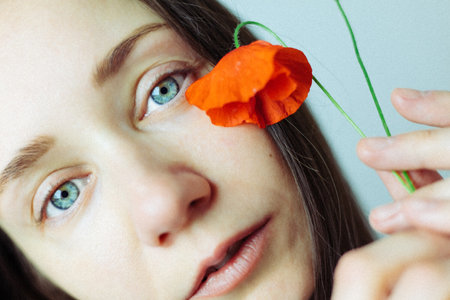 A young woman holds a red poppy flower near her face, beautiful blue eyes and soft features. A sense of delicate beauty and natural female charm.の写真素材