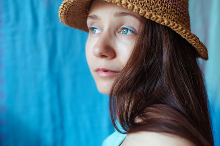 A serene young woman with blue eyeliner wearing a straw hat and looking off into the distance with a thoughtful expression against a blue backdrop.の写真素材