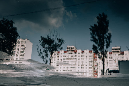 A serene reflection of a building and trees in a puddle on the street. The image has a moody...