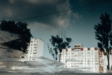 A serene reflection of a building and trees in a puddle on the street. The image has a moody atmosphere. Reality reversed. City street after the rain.の写真素材