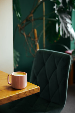 A pink coffee mug sits on a wooden table in a stylish, green-themed cafe setting. A pink mug of coffee rests on a wooden table, set against a backdrop of a green chair and leafy plants.の写真素材