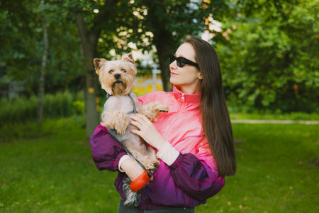 A woman and her dog enjoy a beautiful day in the park together. A stylish girl in black sunglasses holds her Yorkshire Terrier doggy in a park. People walking pets outdoors. Urban dwellers in a park.の写真素材