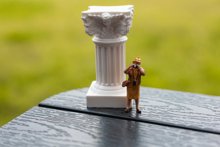 Miniature photographer captures a picture while travels. An old woman figure photographing standing by a classical white pillar, column outdoors. An aging person traveling, taking pictures on a cameraの写真素材
