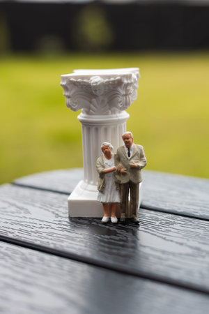 A miniature elderly couple pose near a decorative white column outdoors Old people - retired man and...