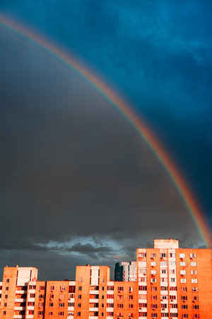 A stunning rainbow stretches across the dramatic blue sky over a building in a residential neighborhood. A vibrant rainbow arcs across a moody, overcast sky above a building.の写真素材