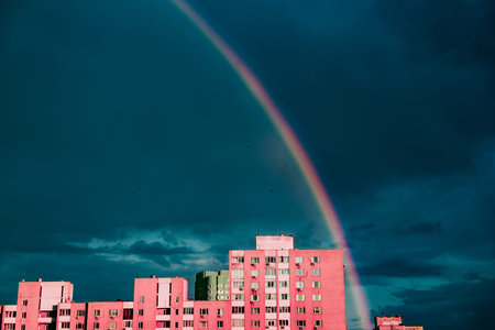A beautiful rainbow appears after a storm over urban buildings. A vibrant rainbow arcs over pink building roof against a dramatic, stormy dark blue sky. Urban landscape. Spring or summer weather.の写真素材