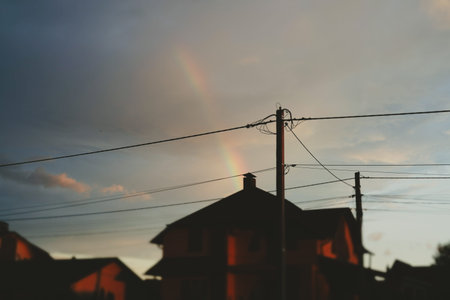 A vibrant rainbow arcs across the dramatic sunset sky over a suburban neighborhood. A stunning rainbow appears above houses and roofs against a colorful evening skyline. The silhouette of buildings.の写真素材