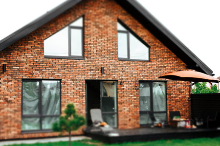 Modern brick house featuring a patio, stylish triangular windows, and outdoor living spaces. A cozy terrace with an orange umbrella and garden furniture. The contemporary architecture exterior design.の写真素材