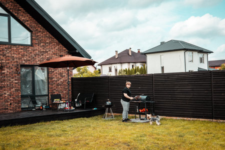 A woman enjoys a barbecue in her backyard with her small dog on a sunny day. A person is grilling food in the backyard. A small doggy is near her. Modern houses and a fence are in the background.の写真素材