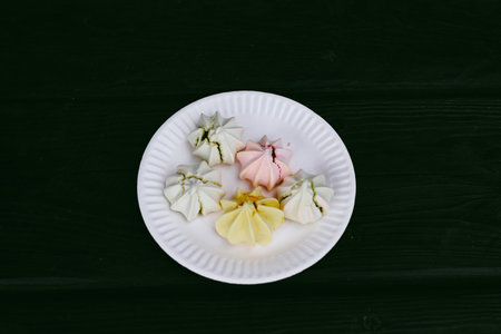 Colorful meringues on a white plate, a delectable dessert. A delightful plate of multicolored meringues sits on a dark brown surface, a sweet treat for children party. Pastel colors Treats and sweetsの写真素材