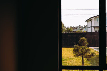 A serene suburban scene is viewed through a window, featuring houses, yards, green grass. A view through a window offers a glimpse of a calm suburban neighborhood with houses, a small tree in a yard.の写真素材