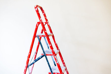 A red step ladder, set against a clean white wall, is ready for use. A red step ladder stands in front of a plain white wall, perfect for home improvement and renovation projects.の写真素材