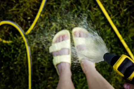 A person sprays water on the feet with a garden hose on a sunny day outdoors. A girl is using a garden hose to spray water onto their feet, wearing light yellow sandals, on a background of green grassの写真素材