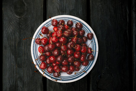 A vibrant image of ripe cherries on a plate set against a dark wooden backdrop. A plate of fresh, juicy cherries sits on a rustic black wooden table, capturing a vibrant, appetizing scene.の写真素材