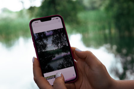 A person views a nature photo on smartphone, showcasing a beautiful lake. A person holding a modern phone that displays a photo of a scenic pond with trees reflected in calm water, blurred background.の写真素材