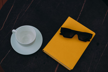 Sunglasses and a yellow book with a cup on a dark, marble table. A cup and saucer, sunglasses, and a...