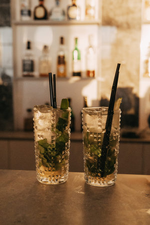 Two beautifully crafted mojitos sit on a bar ready to be enjoyed. Fresh mint, lime and straws make...