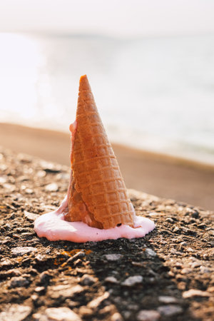 Fallen ice cream cone with spilled pink ice cream on a textured concrete surface. An ice cream cone, tipped over on a rough stone surface with spilled ice cream, and a blurred sea on the background.の写真素材