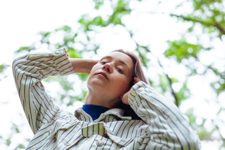 A young woman in a striped shirt looks up with hands behind her head, surrounded by the soft green...