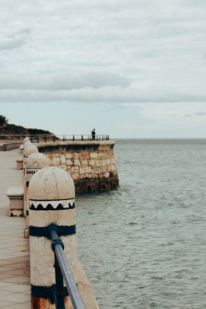 Coastal scene featuring a stone balustrade overlooking the ocean with a person fishing. A scenic...