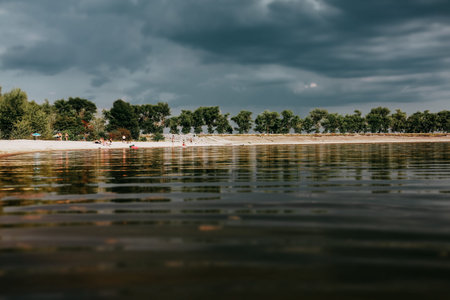 A tranquil lake scene featuring a sandy beach under a dramatic cloudy sky. A serene lake scene with...